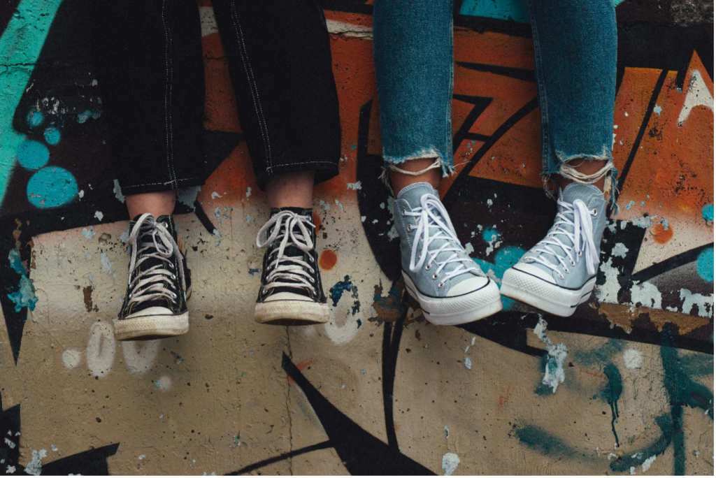 Two people wearing Converse shoes hanging out as their feet dangle against a graffiti wall.